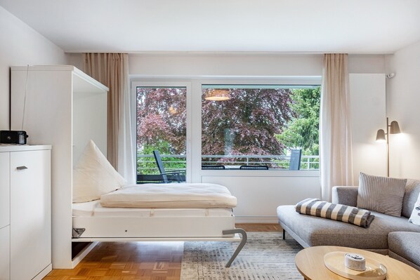 A modern room with a convertible bed, a sofa, a coffee table, and a large window with a view of trees.