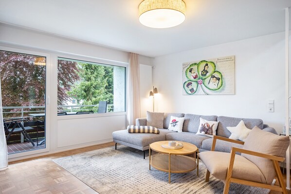 Una sala de estar moderna con un sofá, un sillón, una mesita redonda y ventanas amplias que dan a un paisaje arbolado.