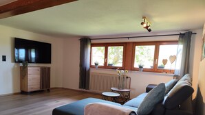 Ein modernes Wohnzimmer mit einer Couch, einem Fernseher, einem Holzschrank, einem kleinen Tisch und einem Fenster mit Vorhängen.