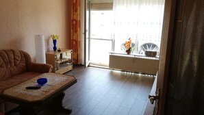 Ein Wohnzimmer mit braunem Sofa, einem Couchtisch mit Fernbedienung, einer Vase mit Blumen und Blick auf einen Balkon mit Stühlen und Heizkörper.