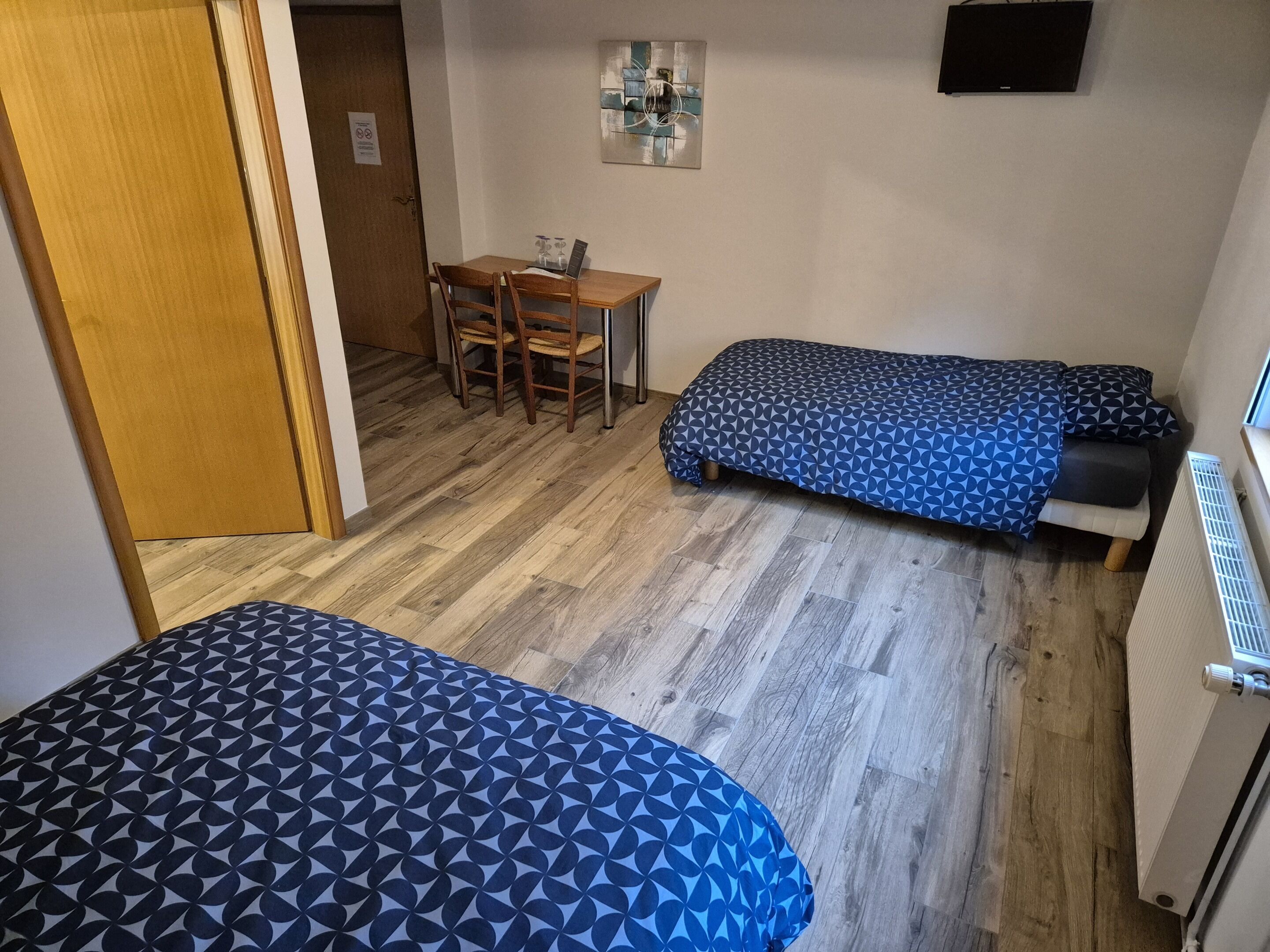 Une petite pièce bien éclairée, avec deux lits, une table en bois et des chaises, ainsi qu’un téléviseur fixé au mur.