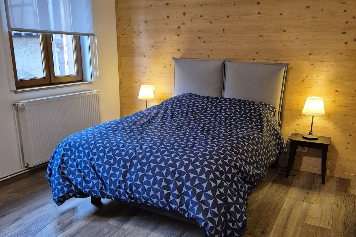 Un dormitorio con una cama, dos lámparas de noche, una ventana con persianas y una pared de madera.