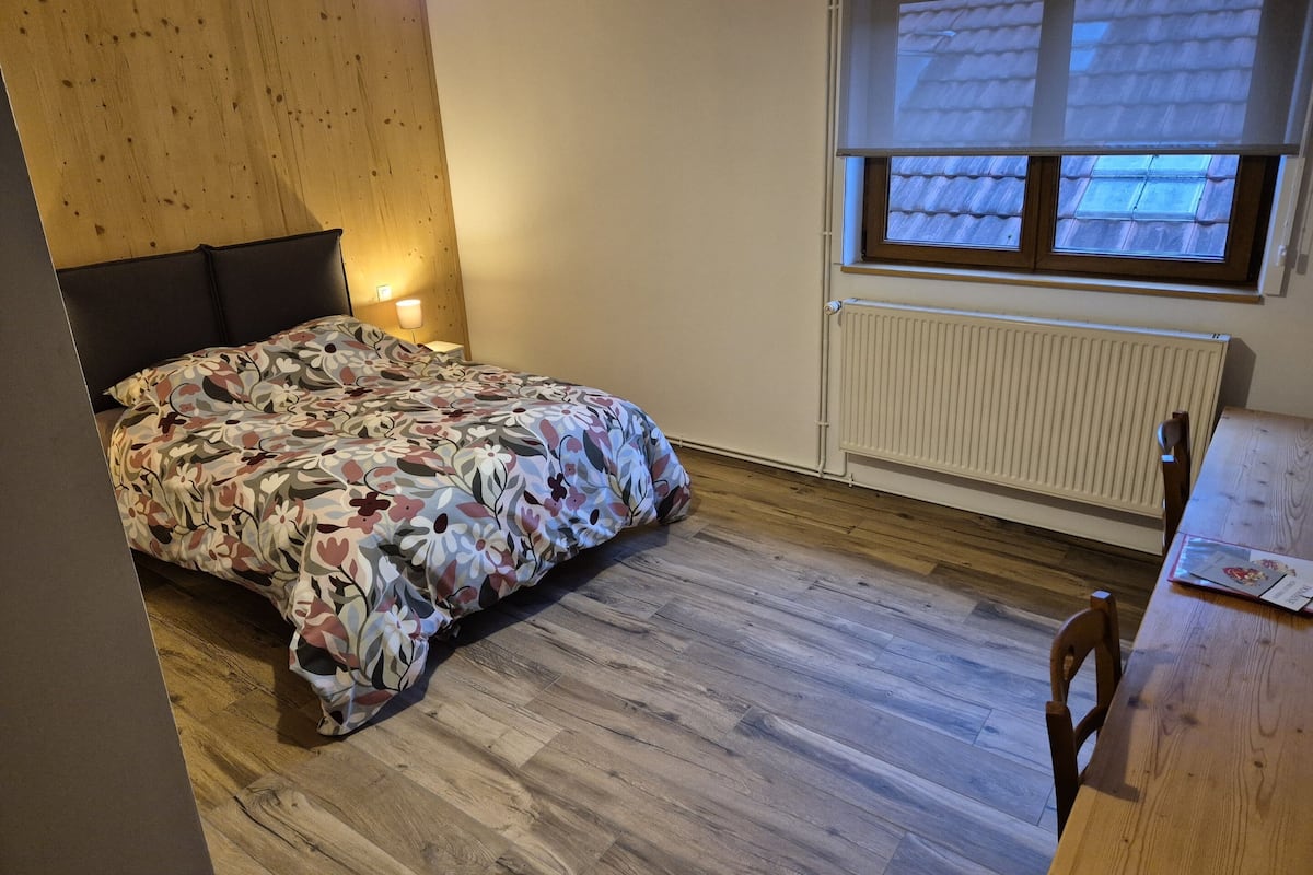 Un dormitorio con cabecera de madera, cama con ropa de cama floral, mesita de noche con lámpara, ventana con vista a un tejado y un radiador.