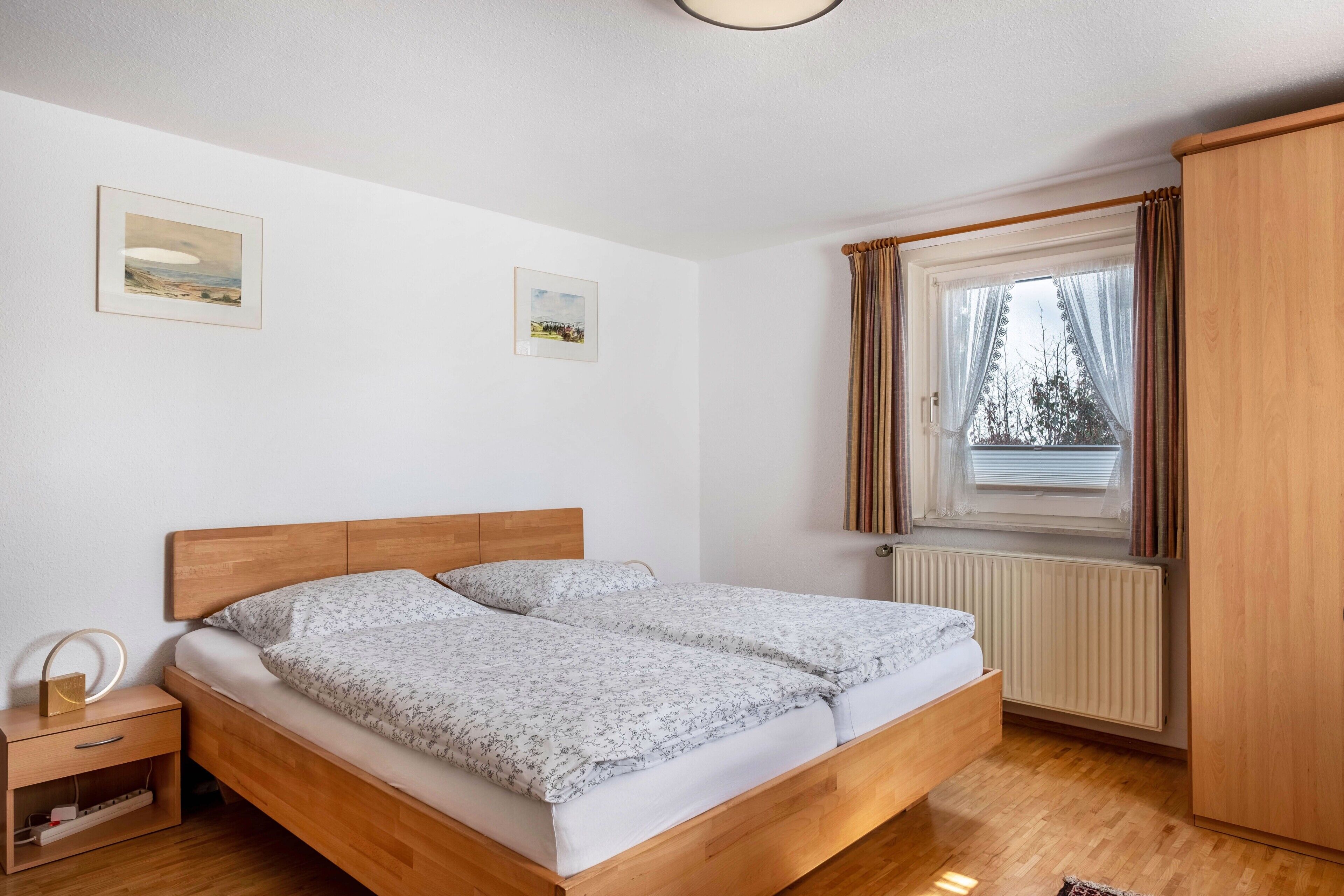 Un dormitorio con cama de madera, una mesita de noche con lámpara, un radiador y una ventana con cortinas.