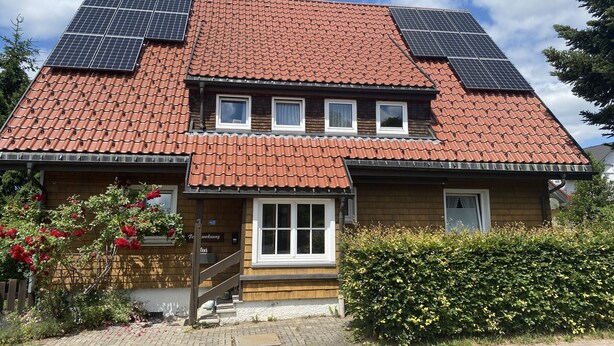 A two-story house with a red-tiled roof, solar panels on the roof, a wooden front porch, and a small garden with red flowers. - Apartment '85qm, 2 Schlafzimmer' with Mountain View, Private Terrace and Wi-Fi (St. Märgen)
