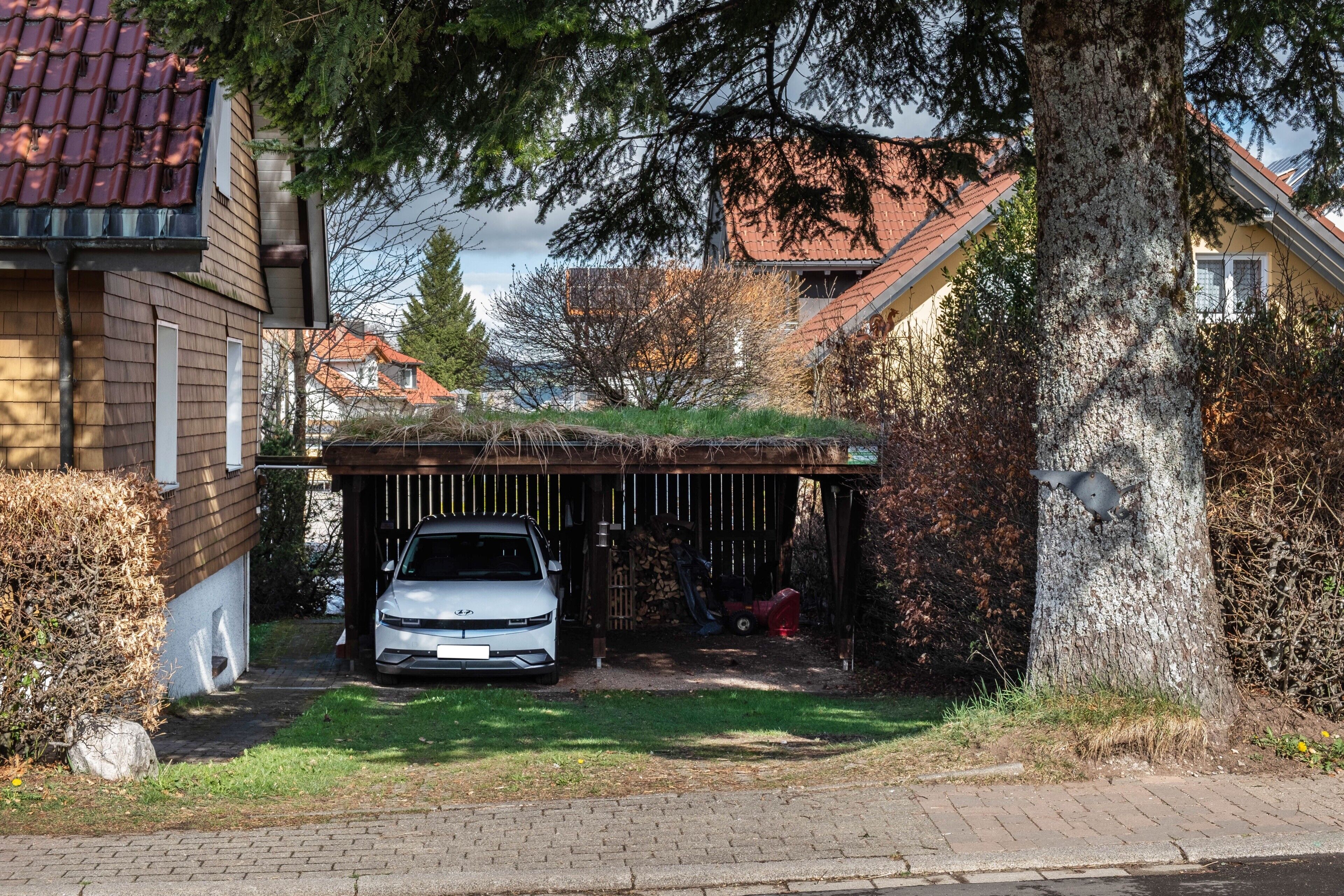 A residential garage with a car parked inside, a tree in the foreground, and houses in the background.