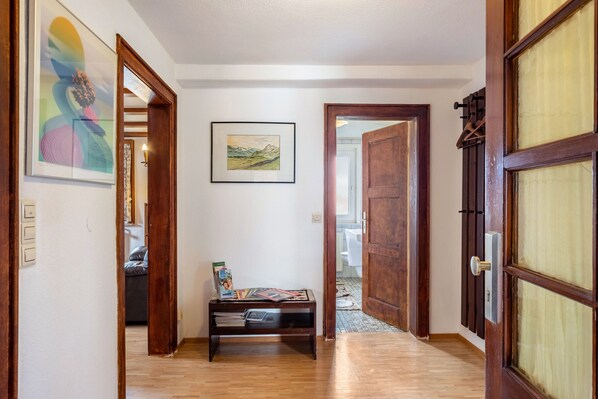 Un pasillo con suelo de madera, un banco con revistas, un cuadro enmarcado con un paisaje y una puerta con panel de vidrio.
