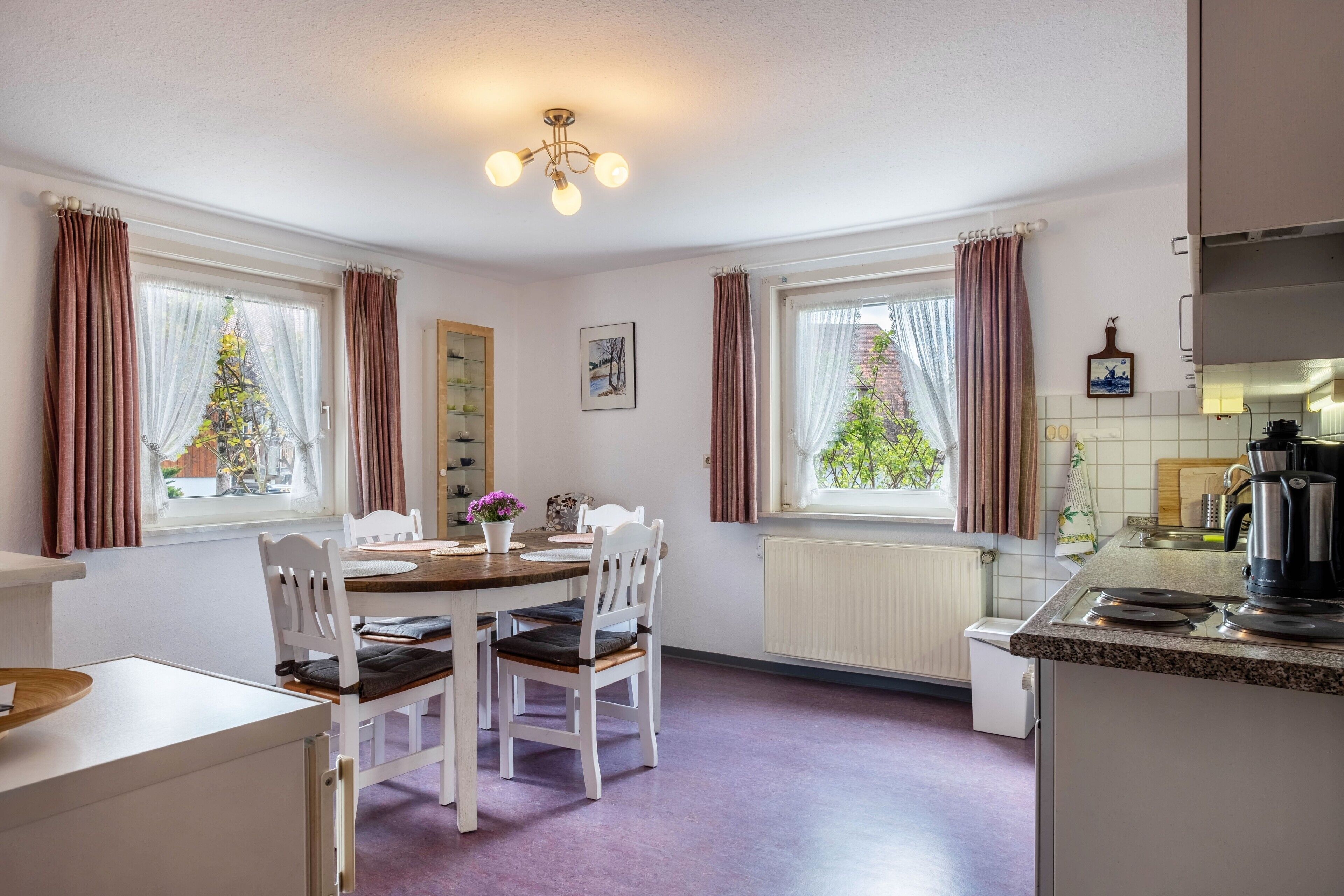 Eine moderne Küche mit weißen Schränken, ein Essbereich mit einem Holztisch und weißen Stühlen sowie große Fenster mit Vorhängen.