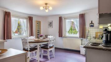 Eine moderne Küche mit weißen Schränken, ein Essbereich mit einem Holztisch und weißen Stühlen sowie große Fenster mit Vorhängen.