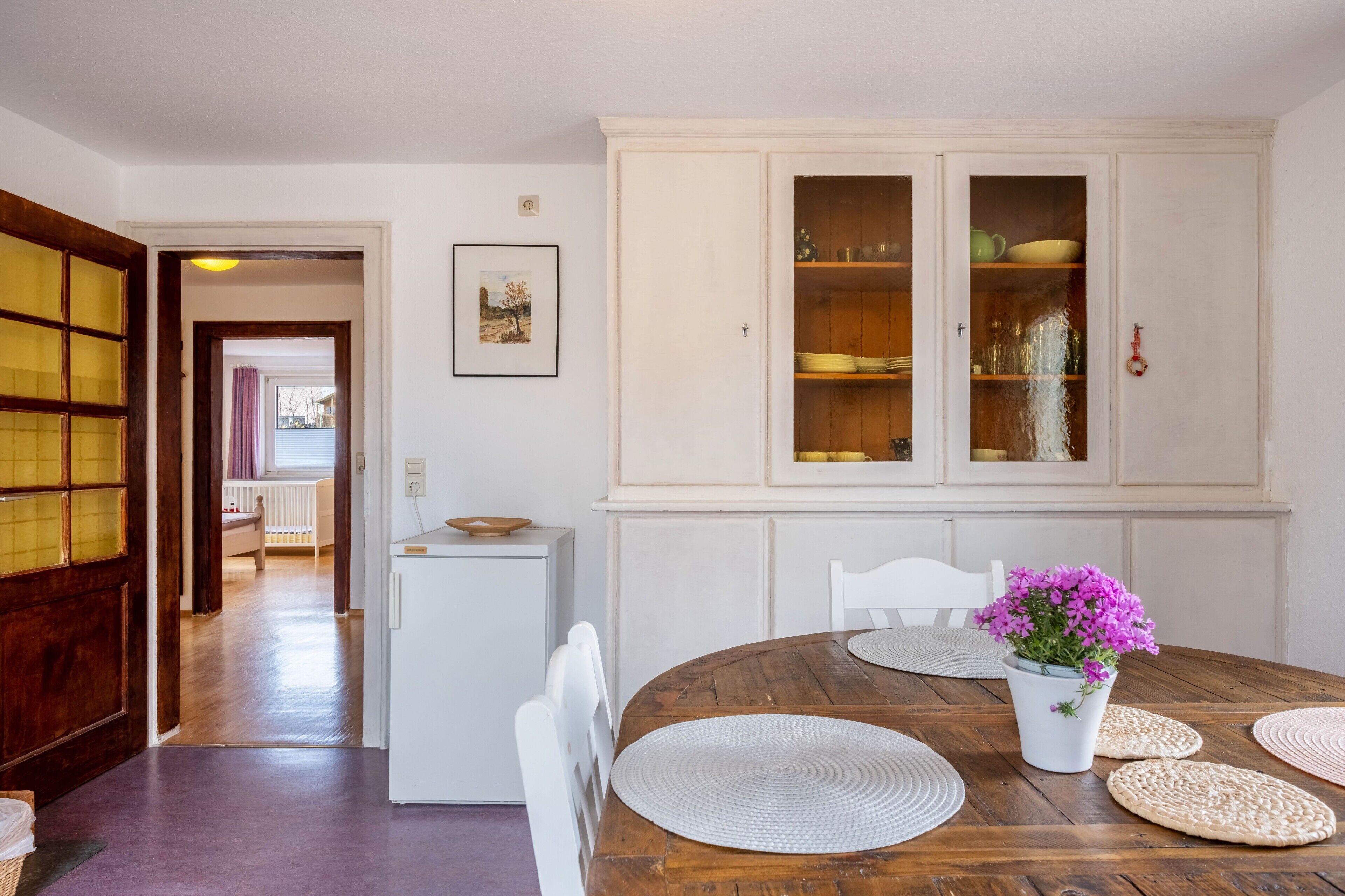 A dining area with a wooden table, white chairs, a white cabinet with glass doors, and a small refrigerator.