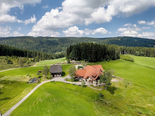 A rural house with a red roof, surrounded by green fields and forested hills.