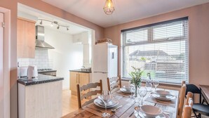 A modern kitchen with a dining area. It includes a kitchen island, a dining table set for four, and a large window with blinds. - Host Stay The Headlands (Coventry)