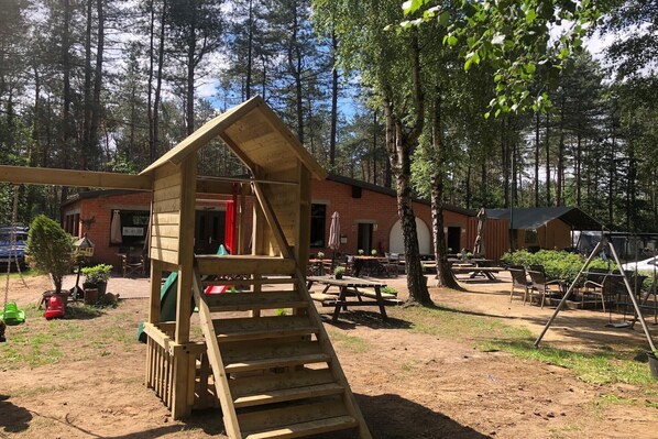 Eine hölzerne Spielkonstruktion in einem Waldgebiet mit Picknicktischen und einem Backsteinhaus im Hintergrund.