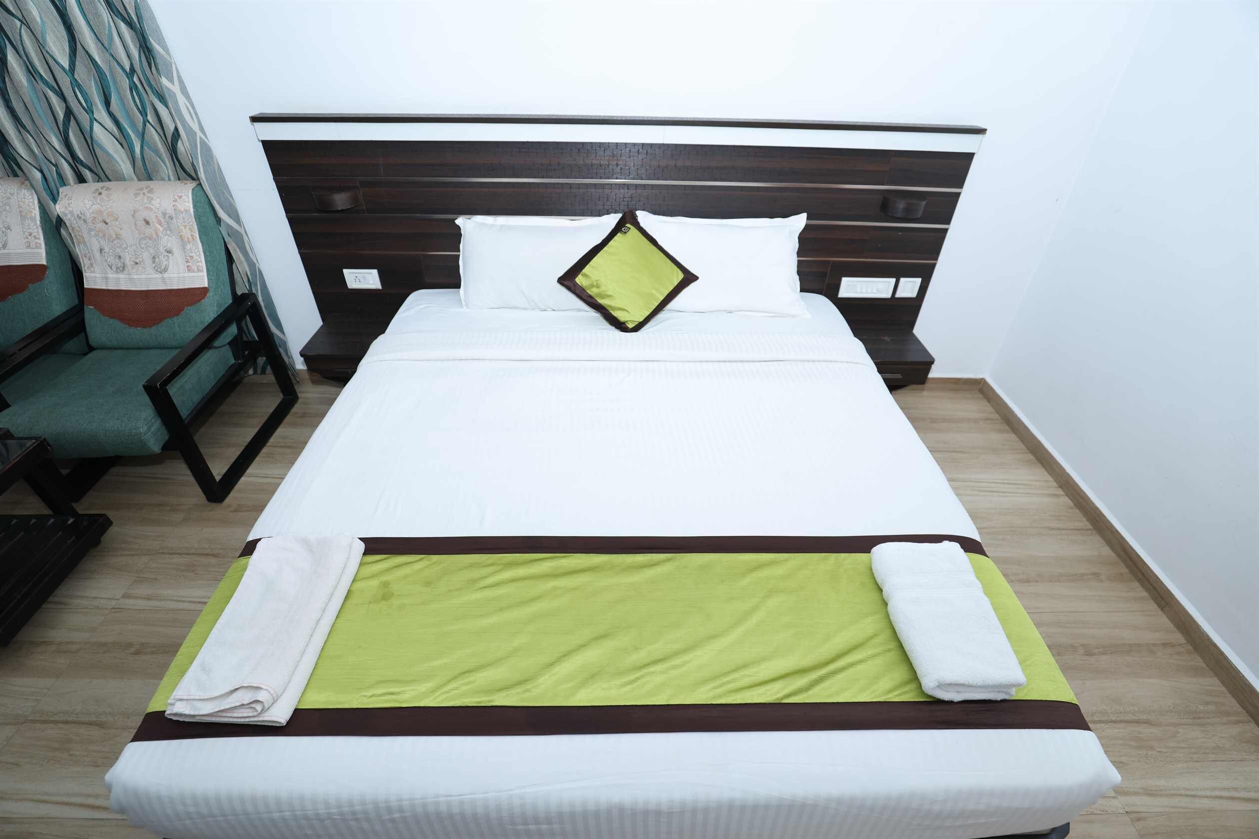 Una cama bien hecha con una colcha verde y marrón, almohadas blancas y una almohada verde con borde negro.