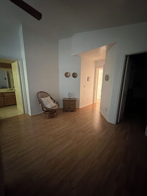 Una habitación amplia con pisos de madera, un sillón de mimbre, una mesita de noche de madera y una puerta que da a otra habitación.