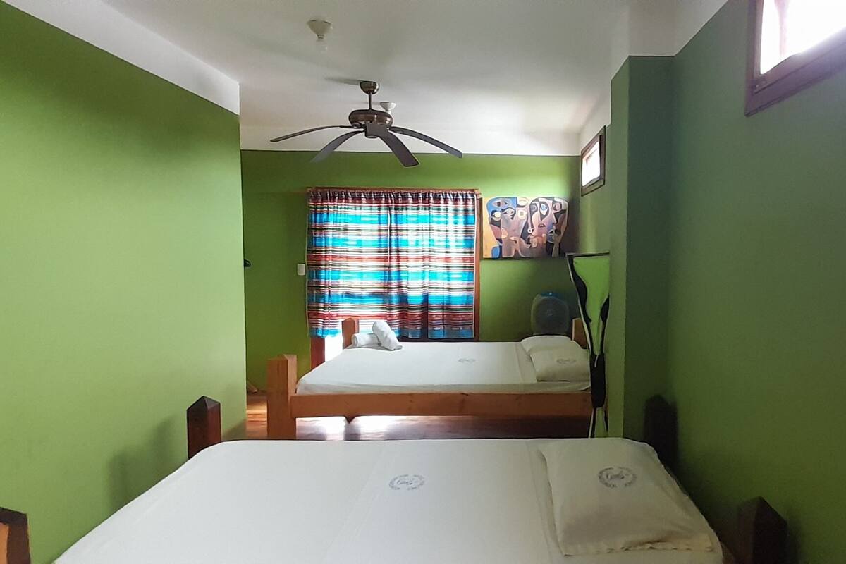 Una habitación con dos camas, un ventilador de techo, una ventana con cortinas y un estante en la pared con un ventilador.