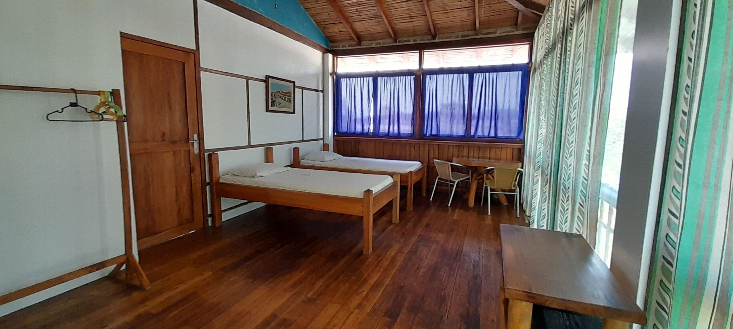 Quarto com duas camas de solteiro, uma mesa de madeira, uma cadeira e uma janela com cortinas.