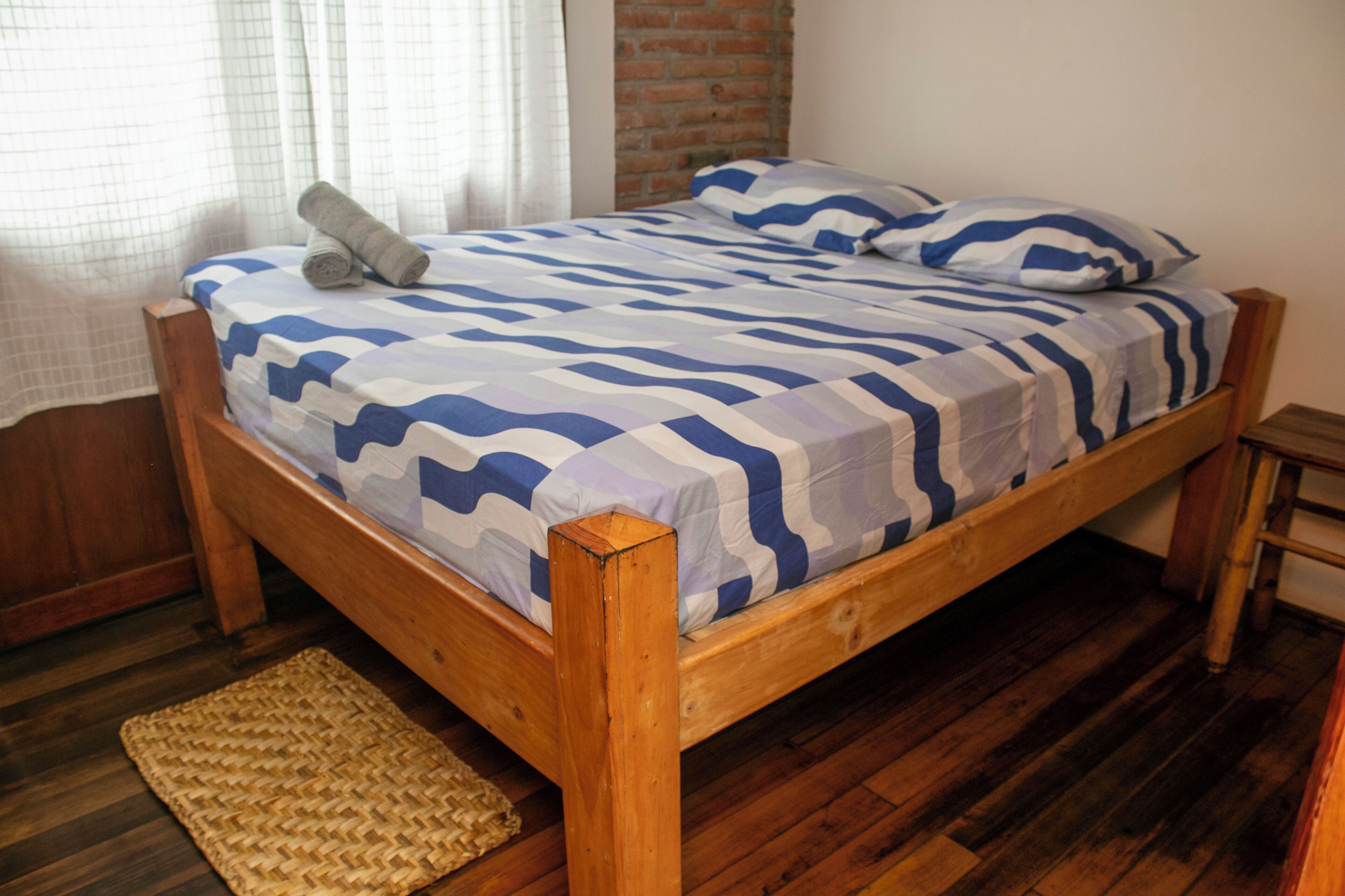 Una cama de madera con colcha y almohadas a rayas azules y blancas, un tapiz tejido en el suelo y una pared de ladrillo detrás de la cama.