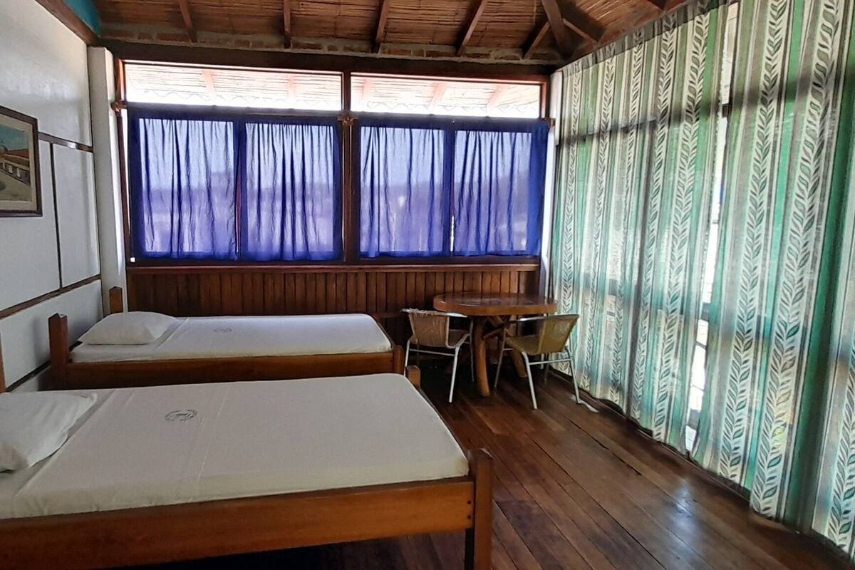 Una habitación con dos camas de madera, una mesita y un ventanal con cortinas.