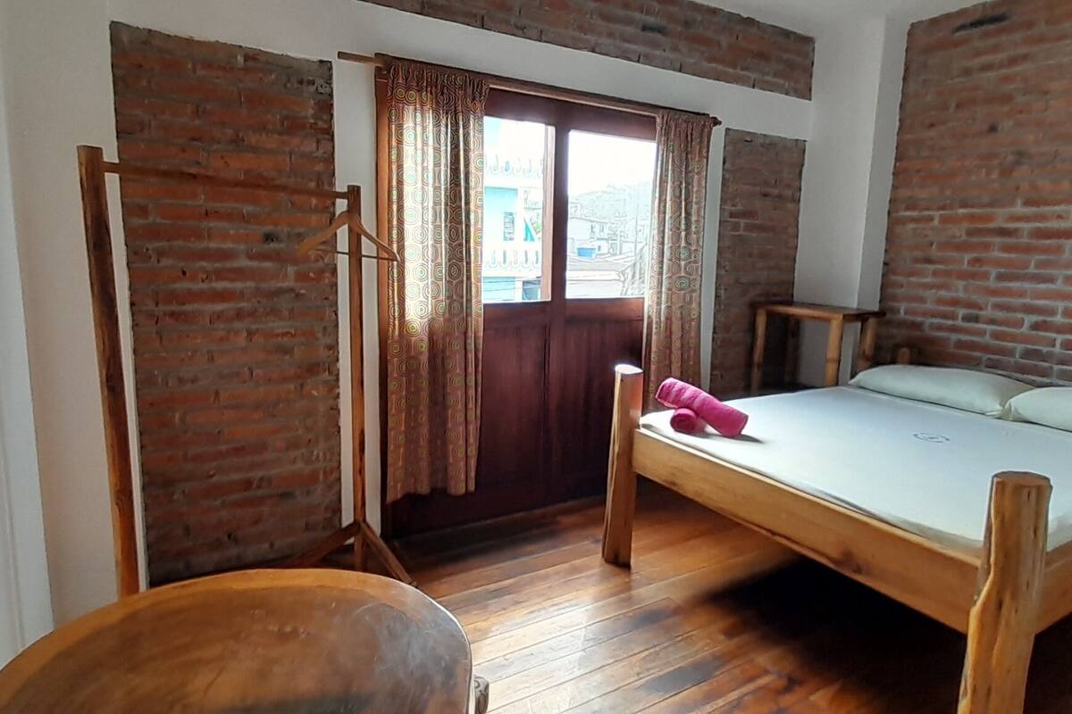 Una habitación con cama, silla y mesa de madera, y una pared de ladrillo.