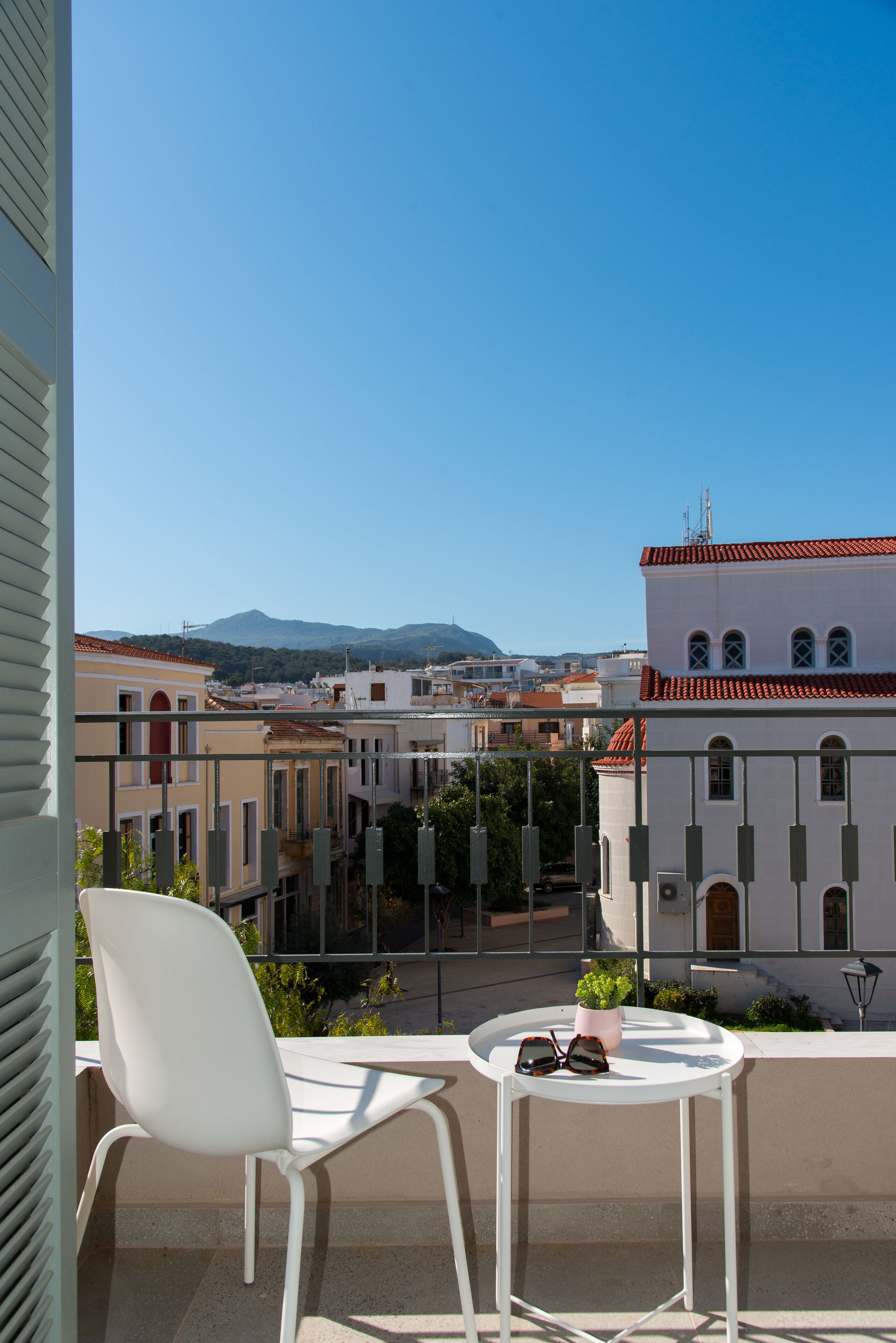 A balcony with white chairs and a small table, overlooking a cityscape with buildings and a mountain in the distance.