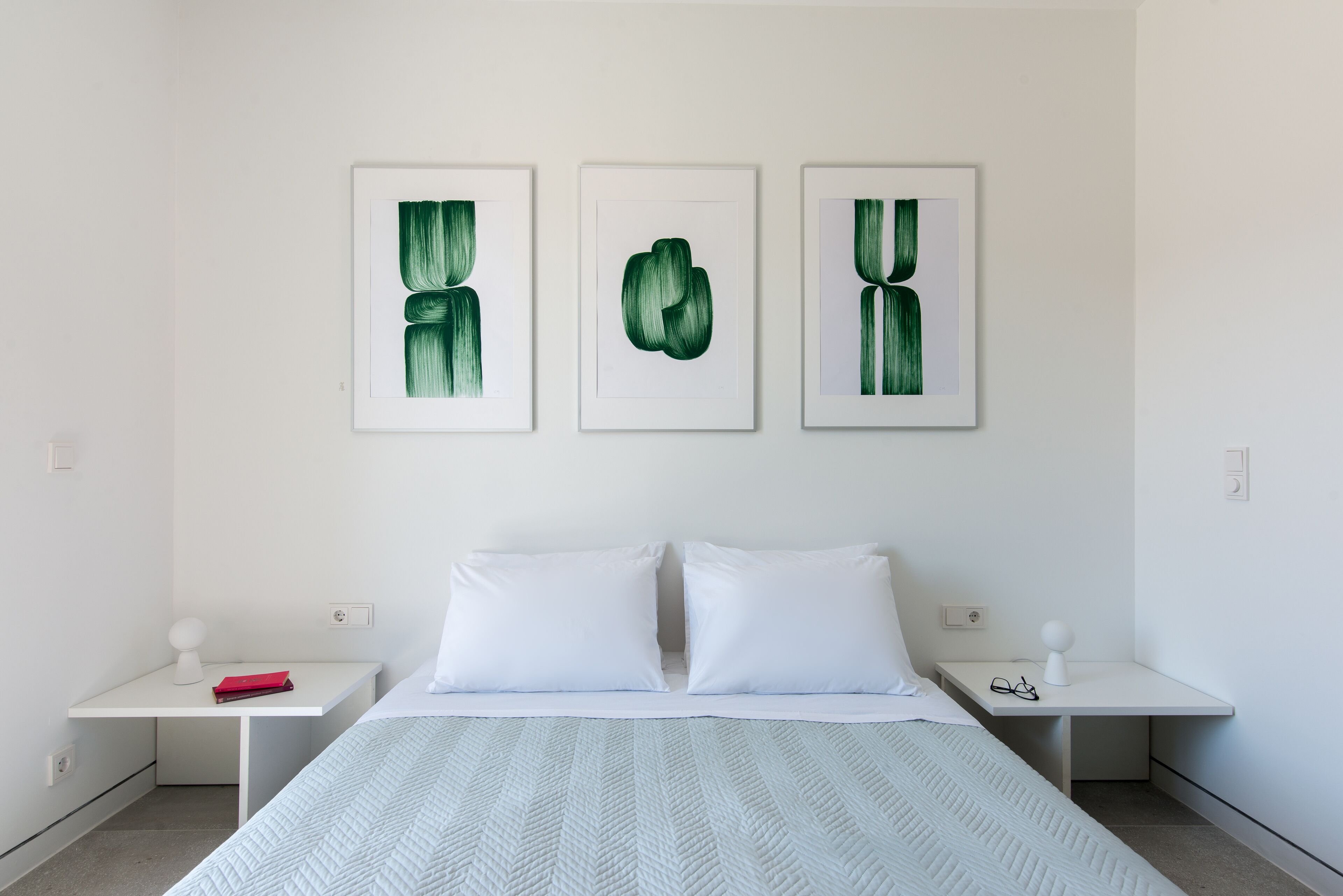 A minimalist bedroom with a bed, white nightstands, and three framed botanical prints on the wall.