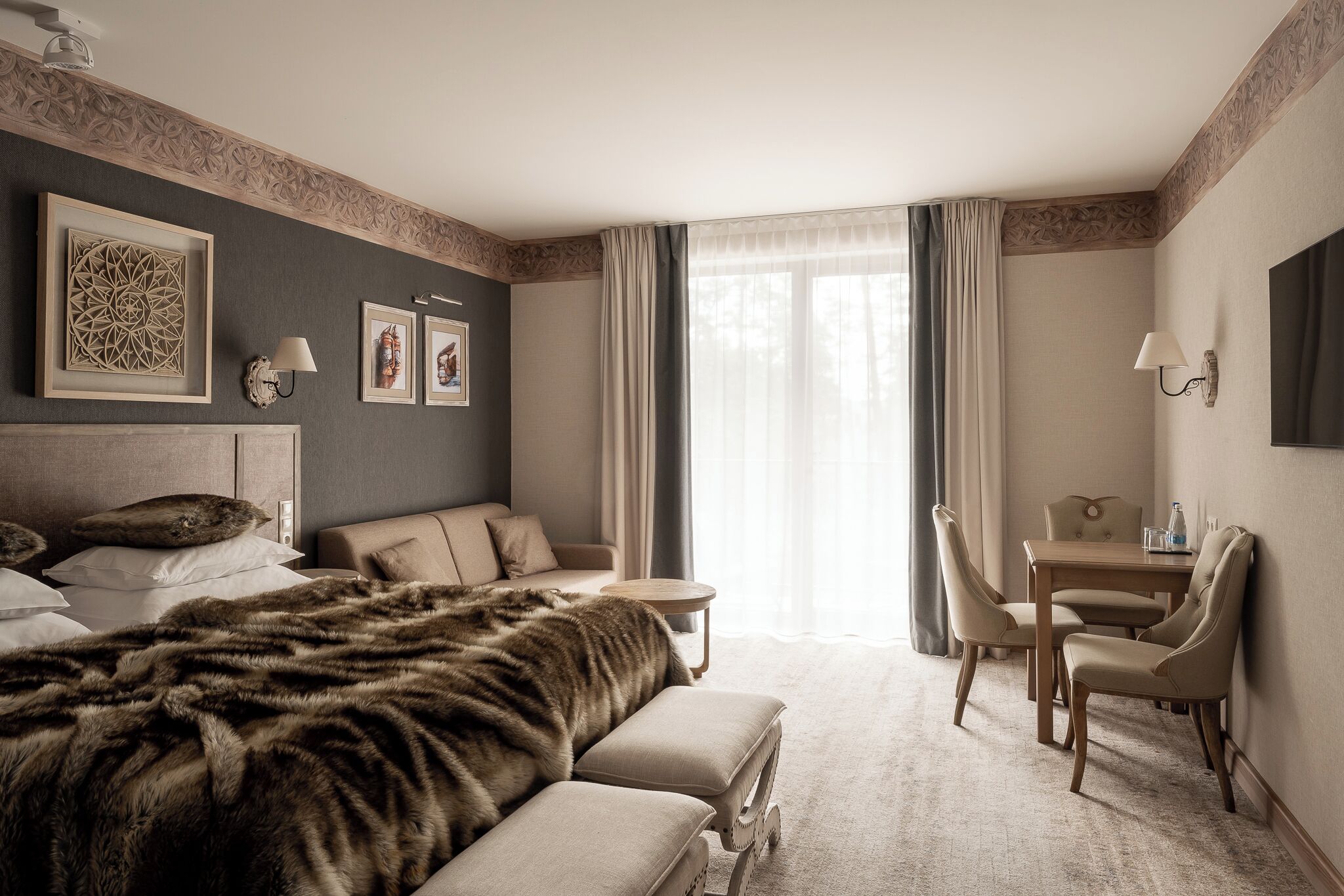 A hotel room with a large bed, a sofa, a dining table with chairs, a TV, and artwork on the walls.