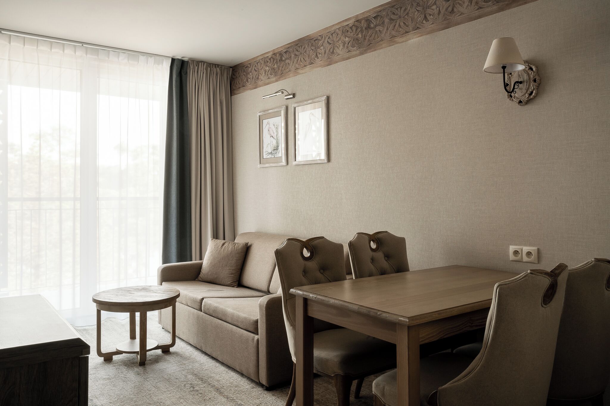 A hotel room with a sofa, a dining table, a small round table, a wall-mounted lamp, and two framed pictures.