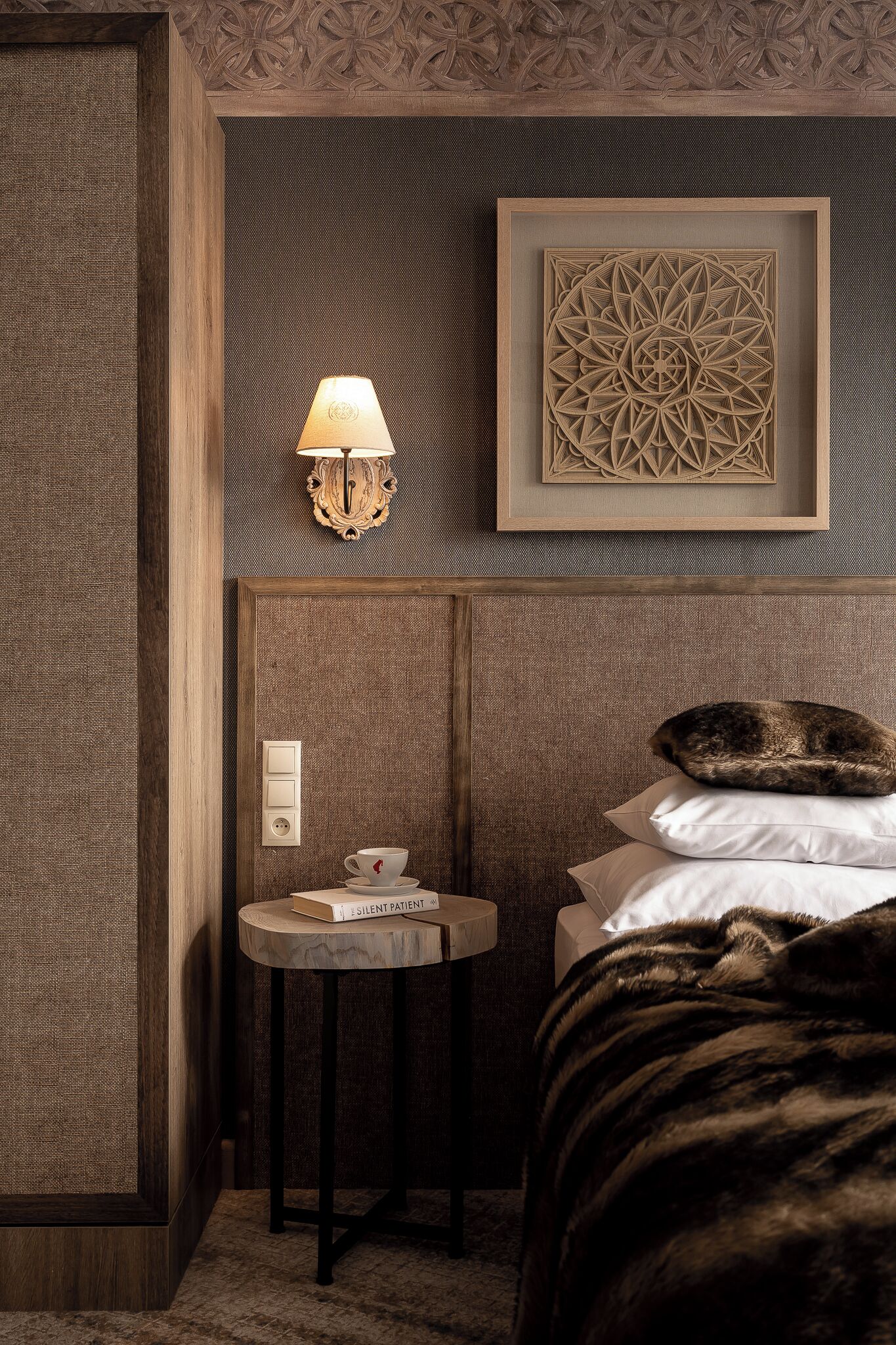 A hotel room with a bed, a bedside table with a lamp, a framed geometric artwork, and a wall switch.