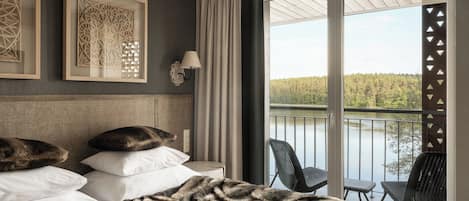 A hotel room with a bed, a nightstand, a lamp, a framed artwork, and a balcony with a view of a forest and water.