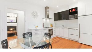 A modern kitchen with white cabinets, a black microwave, and a stainless steel range hood. There is a glass dining table with chairs. - Bayside Home Away From Home (Brighton)