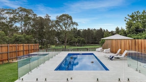 A rectangular swimming pool with a glass enclosure, surrounded by a wooden fence and a grassy area.