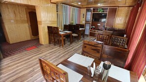 Un espace repas au sol en bois, avec des tables et des chaises, un téléviseur et un meuble sur lequel est posée une plante.