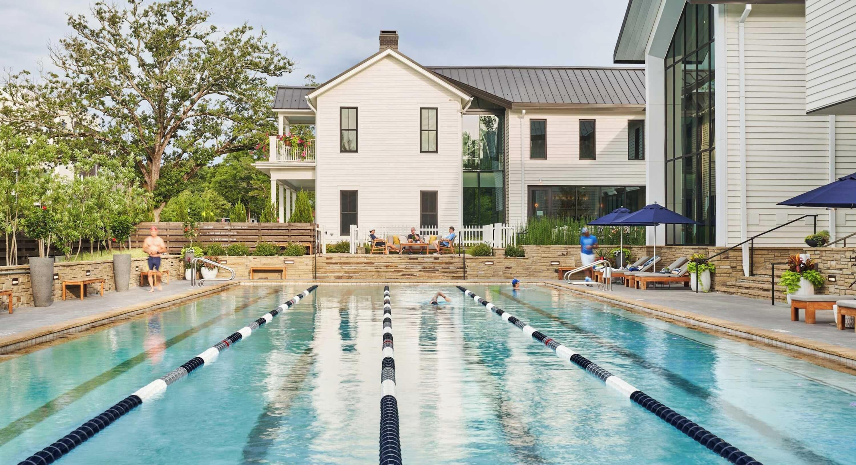 A swimming pool with lane dividers, surrounded by lounge chairs and umbrellas, with a building in the background.