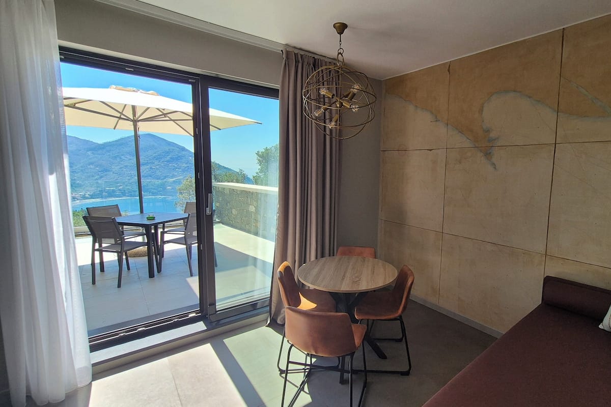 A modern room with a large sliding glass door leading to an outdoor terrace with a table and chairs, a round wooden table, a cushioned sofa, and a decorative ceiling light.