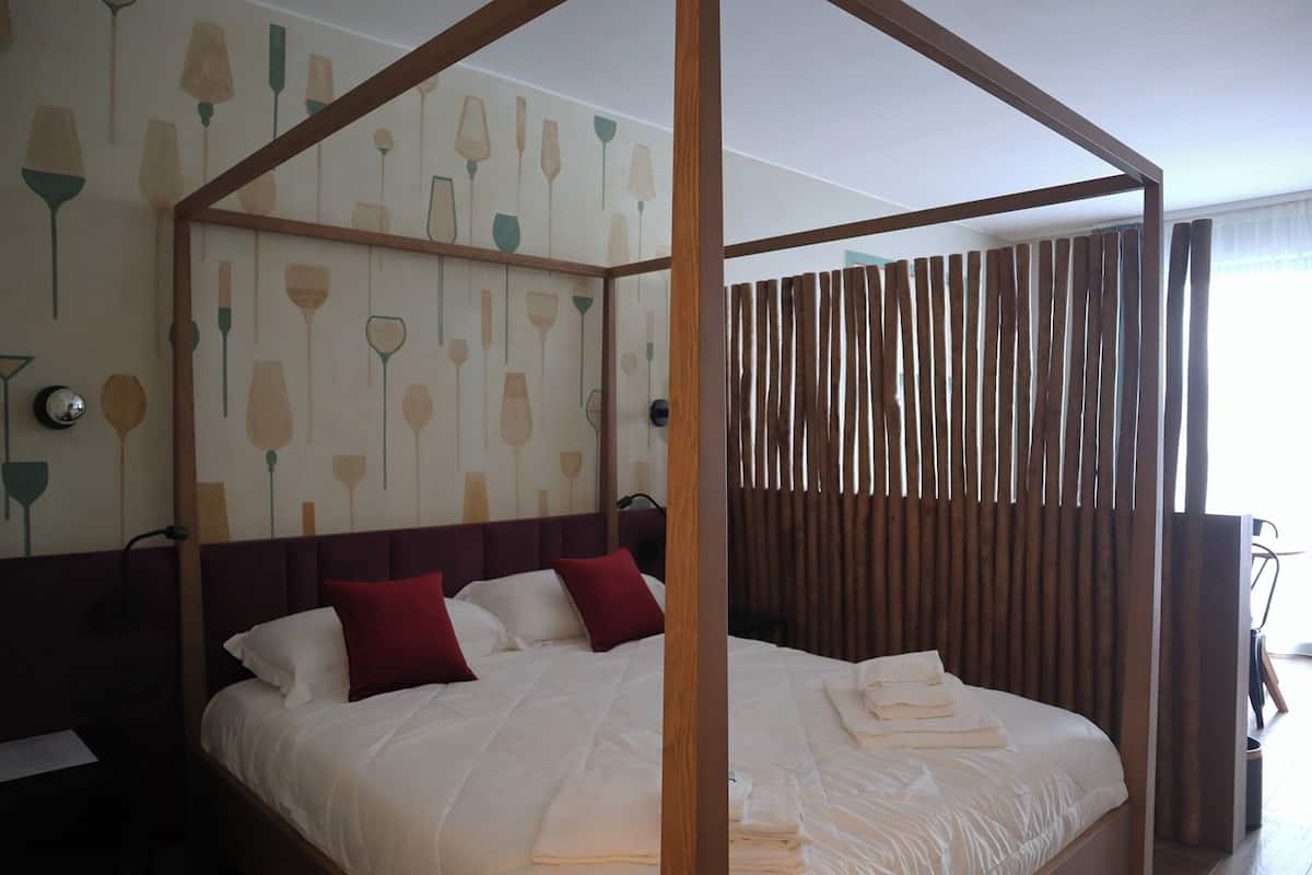 A four-poster bed with white linens and red pillows, a wooden headboard, and a wall with a floral wallpaper design.