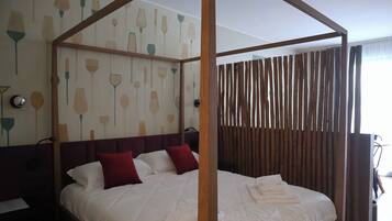 Una cama con dosel tapizada con sábanas blancas y almohadas rojas, cabecera de madera y pared con diseño de papel tapiz floral.