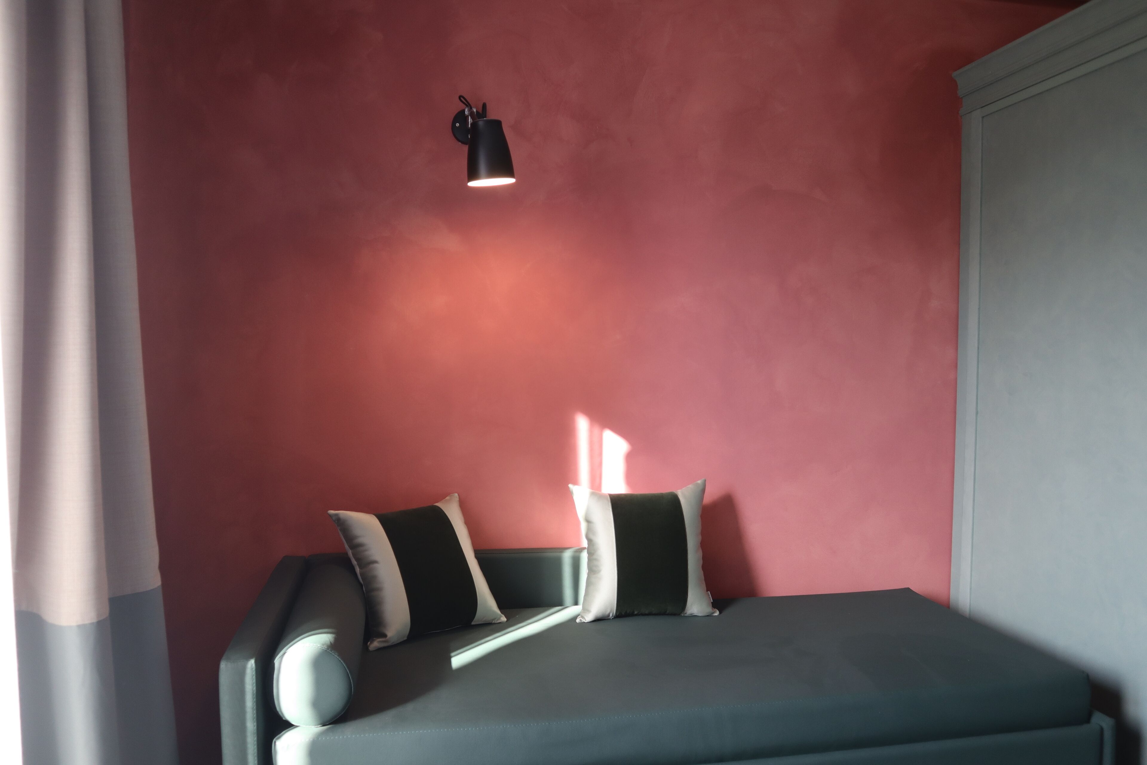 A bed with a grey mattress and white pillows against a red wall, with a black wall-mounted lamp above.