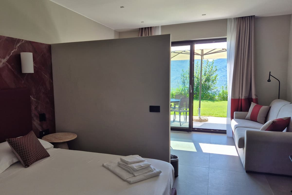A hotel room with a bed, a sofa, a small table, and a sliding glass door leading to an outdoor area with a view of greenery and a mountain.