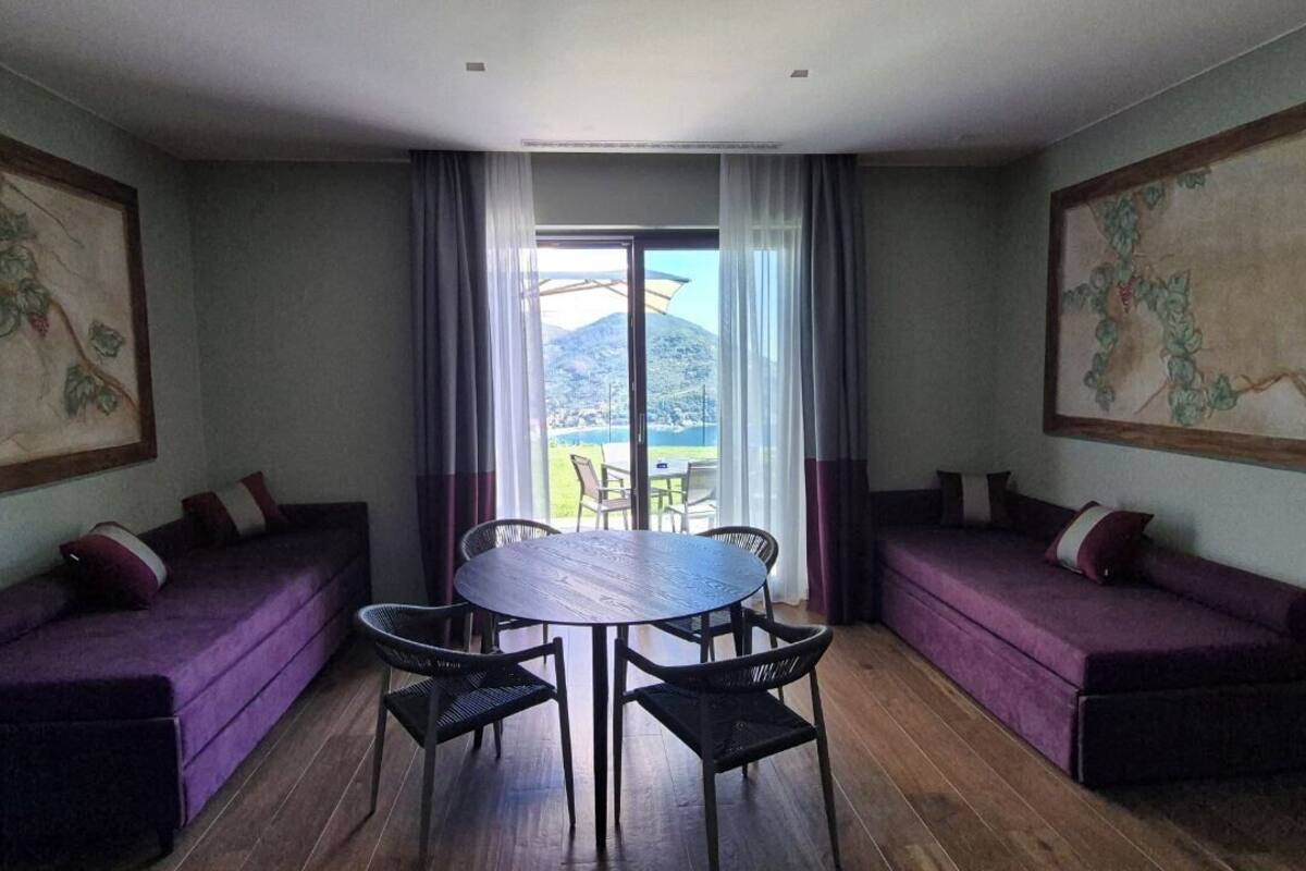 A room with two purple sofas, a round table, and chairs. There is a large window with curtains, and paintings on the walls.