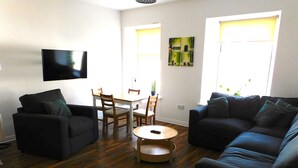 A living room with two sofas, a dining table with chairs, a TV mounted on the wall, and a window with blinds.