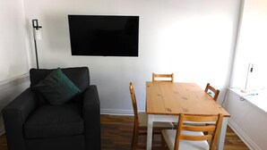 A living room with a black sofa, a wooden dining table with four chairs, and a flat-screen TV mounted on the wall.