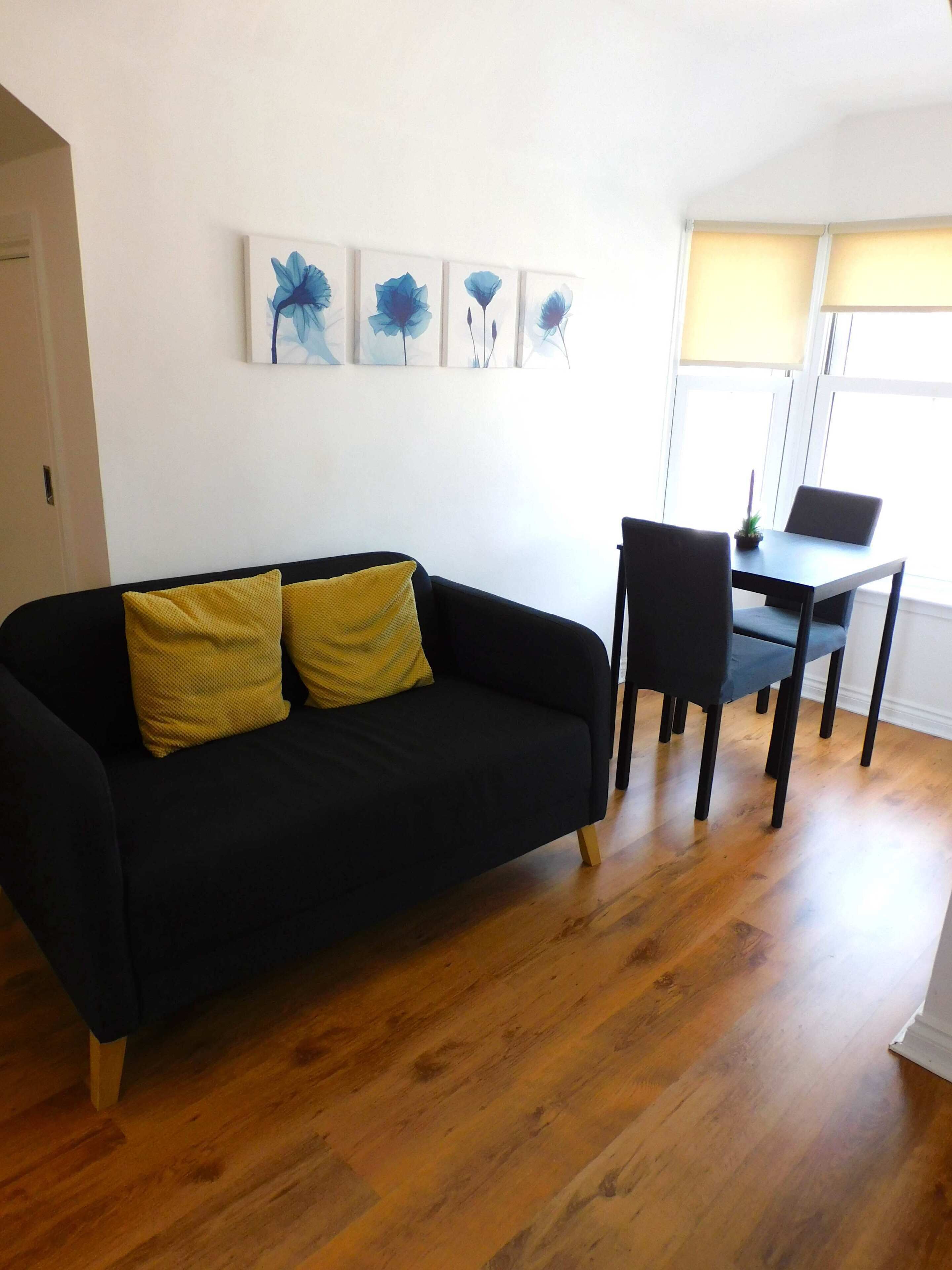 Een woonkamer met een zwarte bank, twee gele kussens, een eettafel met stoelen en een raam met jaloezieën.