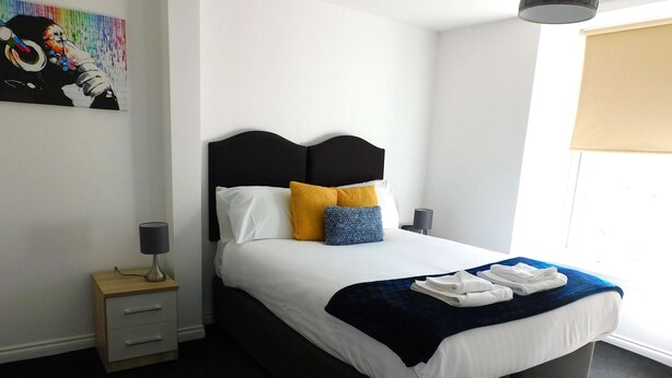 A neatly made bed with white and blue bedding, a dark headboard, and a bedside table with a lamp.