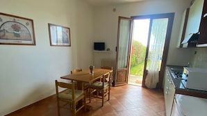 Ein Zimmer mit Esstisch, Stühlen, Fernseher, Küchenzeile und einer Glasfenstertür, die in einen Garten führt.