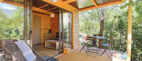 Una terraza de madera con un ventanal que da a una habitación, una mesa con sillas y un sombrero sobre una silla.