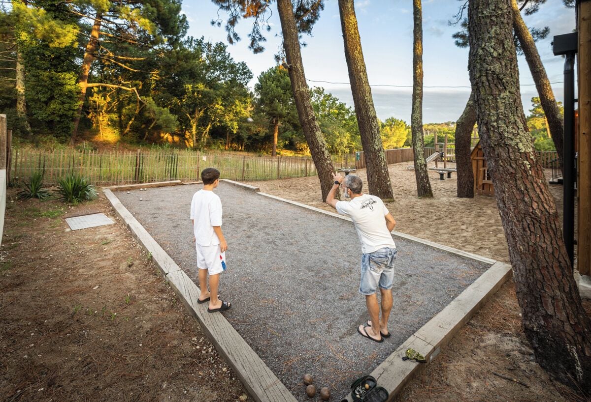 Two people playing shuffleboard on a gravel court with trees and a wooden structure in the background.