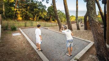 Two people playing shuffleboard on a gravel court with trees and a wooden structure in the background.