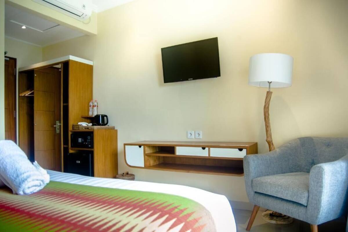 A hotel room with a bed, a TV mounted on the wall, a chair, a desk with a lamp, and a wardrobe.