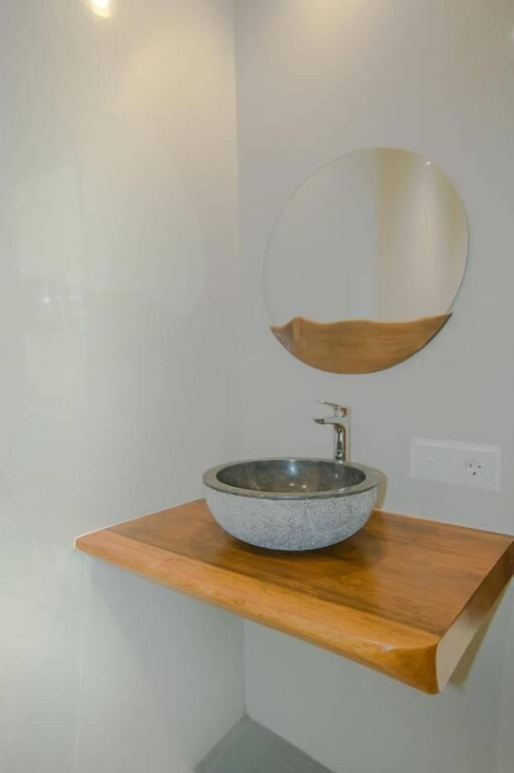 A bathroom with a stone sink on a wooden shelf, a wooden bowl-shaped mirror, and a white wall.